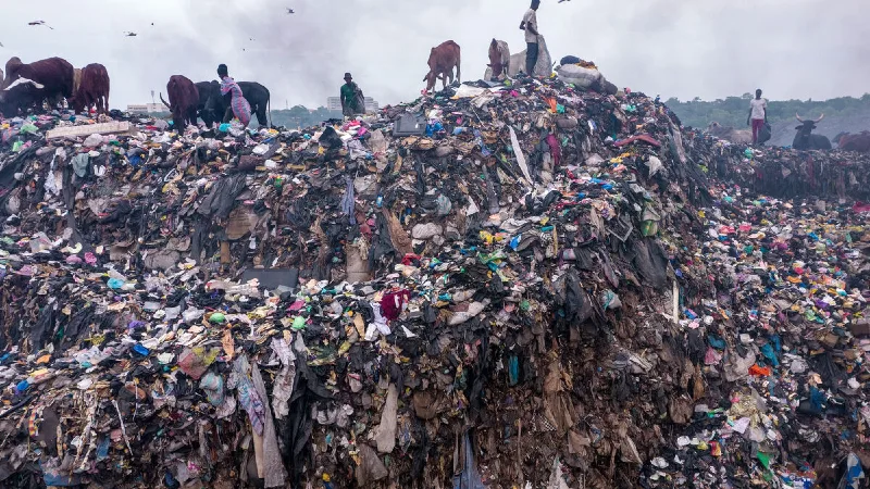 People in Ghana standing on top of a pile of garbage made of discarded fast fashion clothing discarded from western nations.