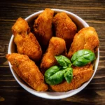 Photo of white bowl with baked breaded alligator nuggets on wood background.